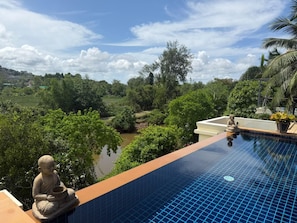 Une piscine ornée d’une statue de Bouddha, entourée d’une végétation luxuriante et offrant une vue sur une rivière.