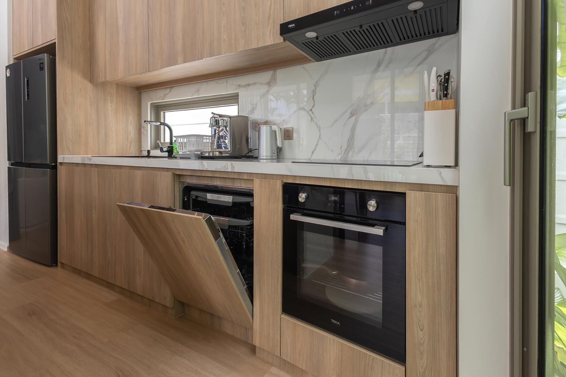 A modern kitchen with wood cabinetry, a built-in oven, a dishwasher, and a stainless steel refrigerator.