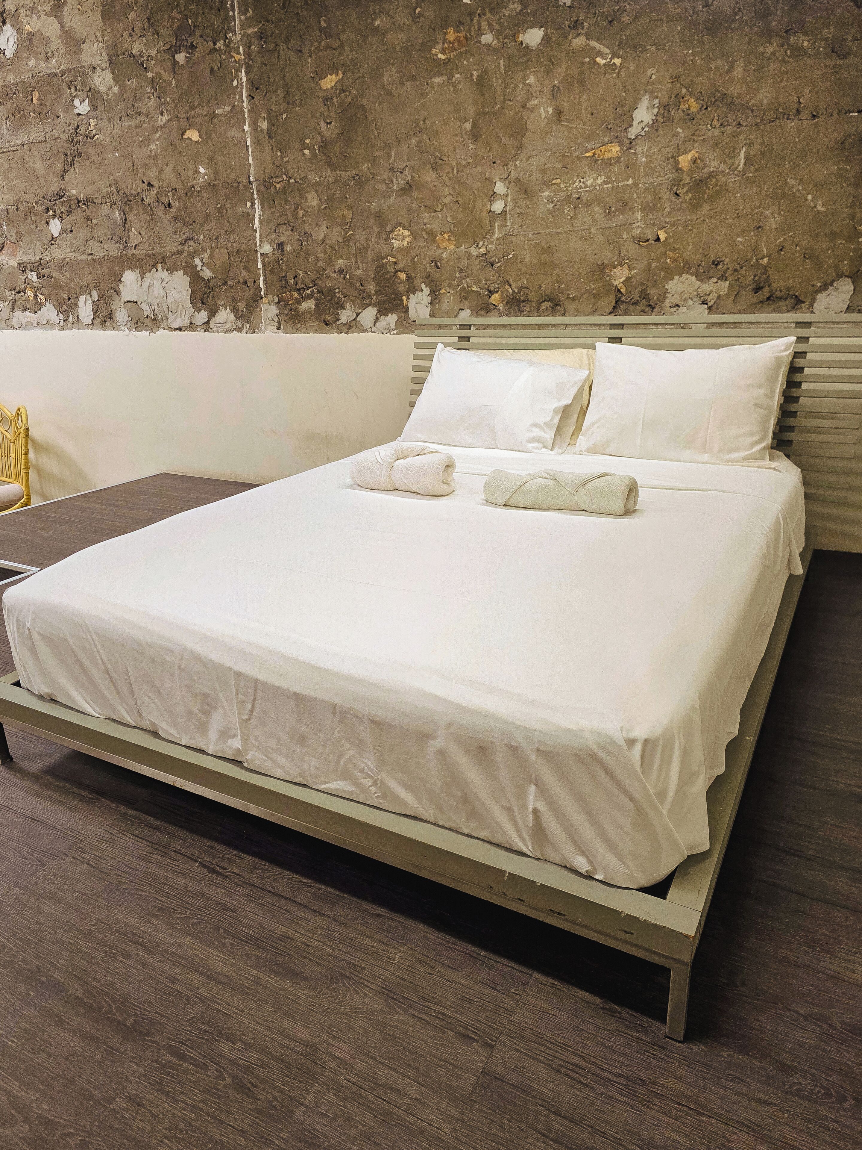 Una cama con sábanas blancas, cabecera de madera y pared lisa con pintura descascarada.