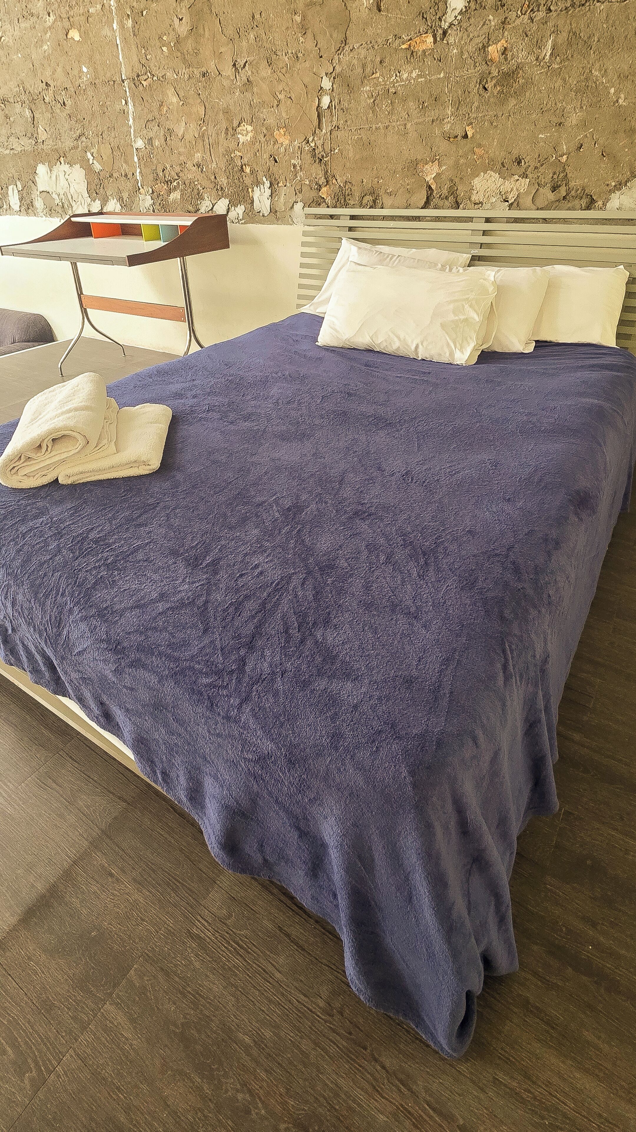 A neatly made bed with a blue blanket and white pillows, a wooden headboard, and a wooden floor.