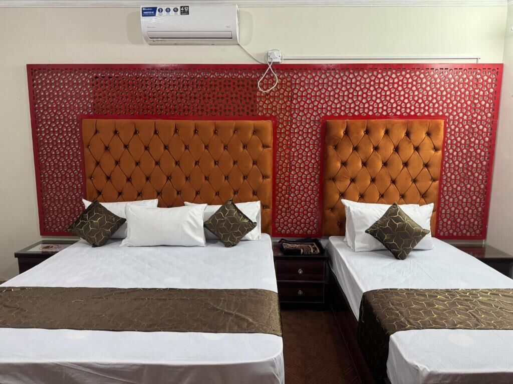 A hotel room with two beds, each with a tufted headboard, white bedding, and decorative pillows. There is a red decorative panel behind the beds, and an air conditioning unit mounted on the wall.