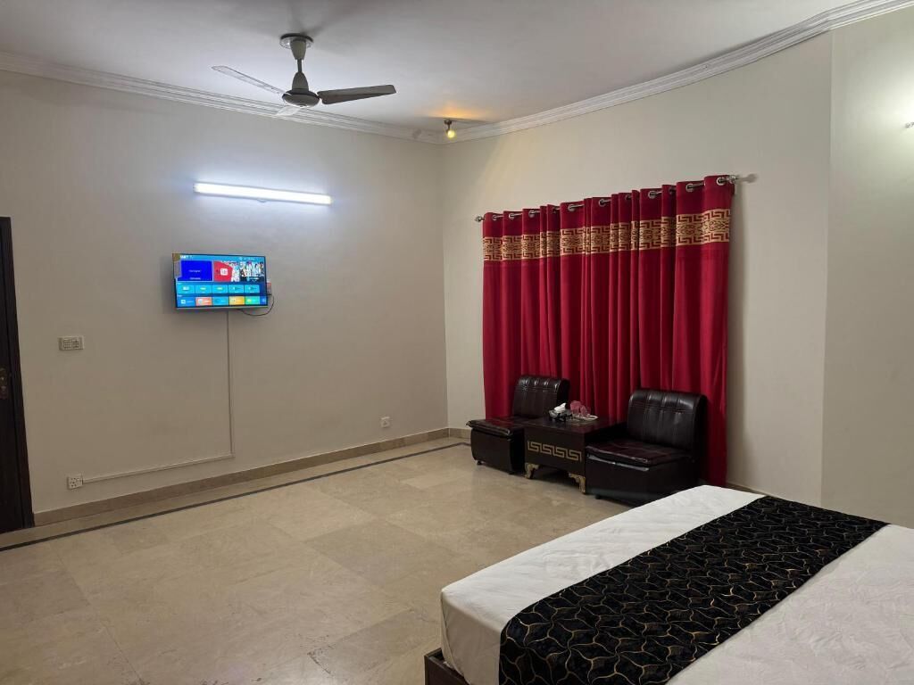 Quarto de hotel com uma cama, uma escrivaninha com cadeira, televisão fixada na parede e ventoinha no teto.