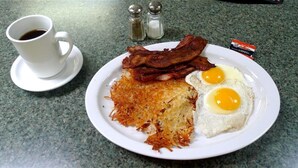 Un plato de desayuno con tocino, huevos y patatas ralladas sobre una mesa, junto a una taza de café y saleros y pimenteros.