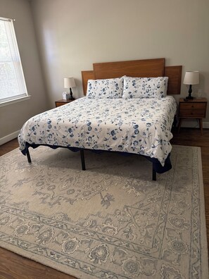 Un dormitorio con una cama, dos mesitas de noche, una ventana con persianas y una alfombra estampada.