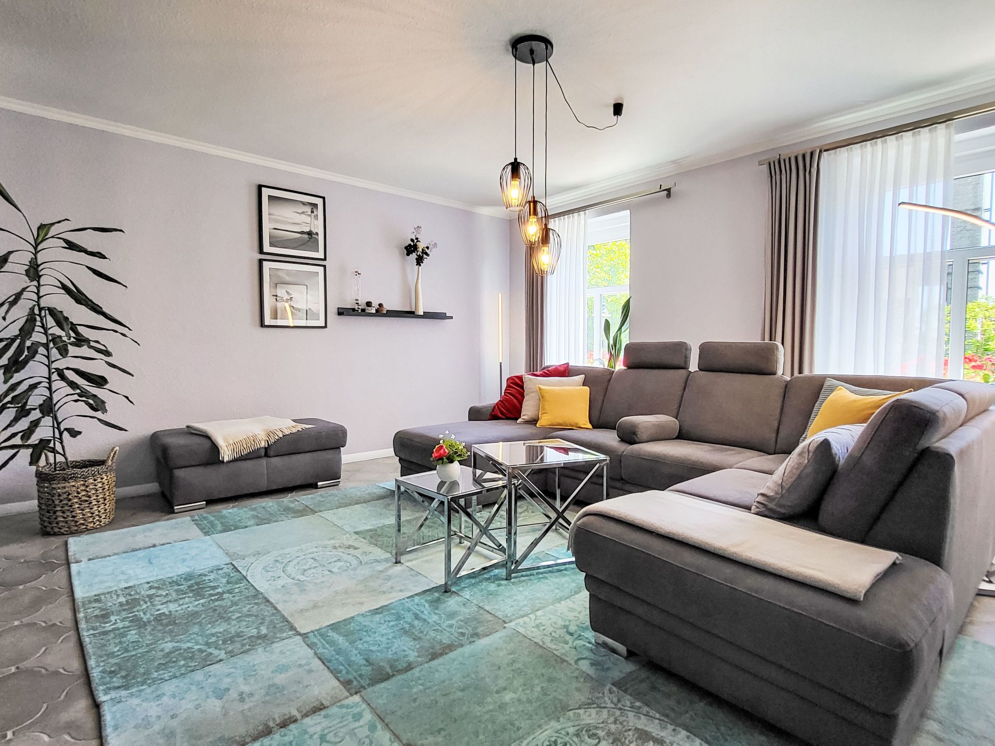En moderne stue med en grå sofa, et sofabord, en stueplante og et stort vindue med gardiner.