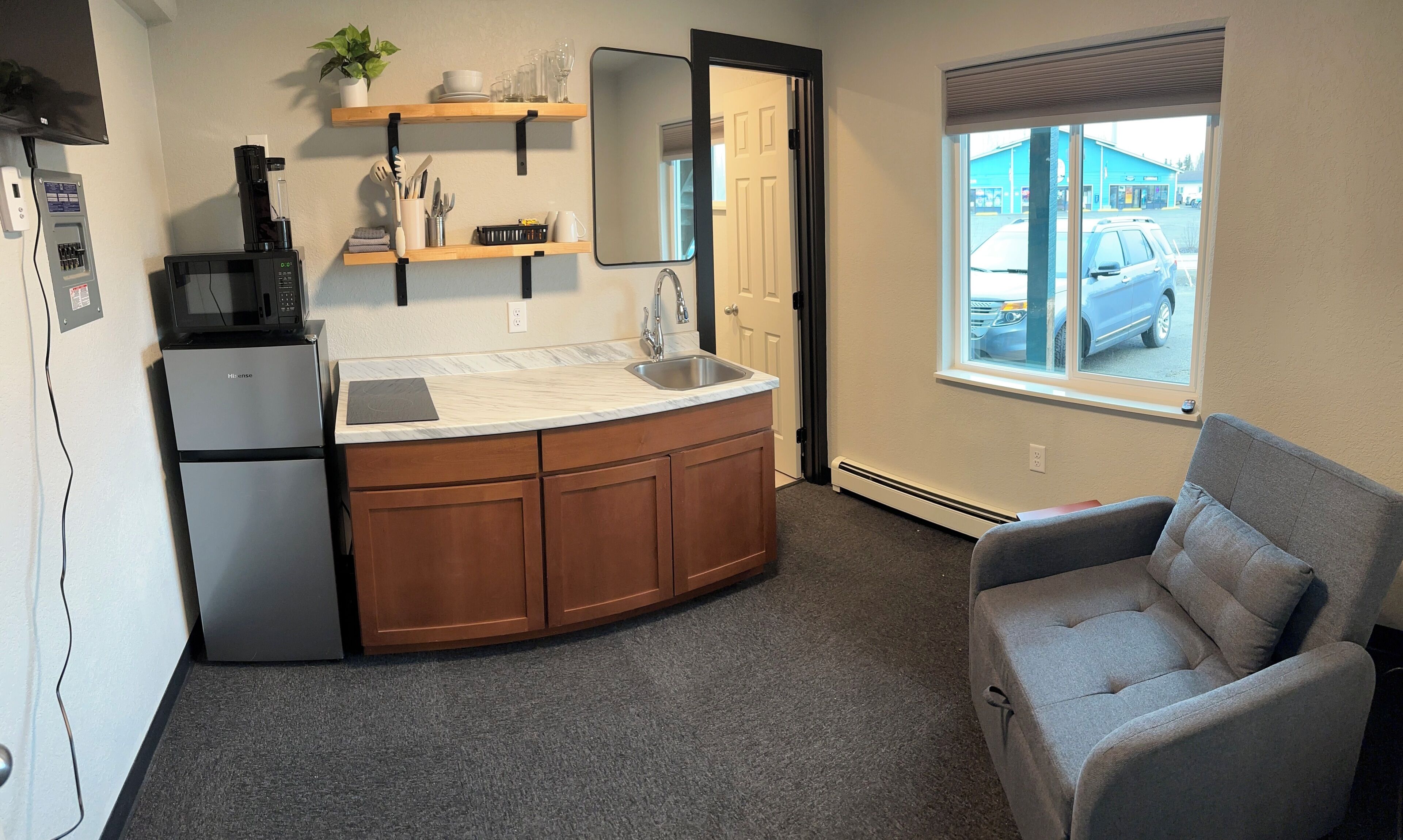 A compact kitchenette with a refrigerator, microwave, and sink, a small seating area with a chair, and a window with a view of the outside.