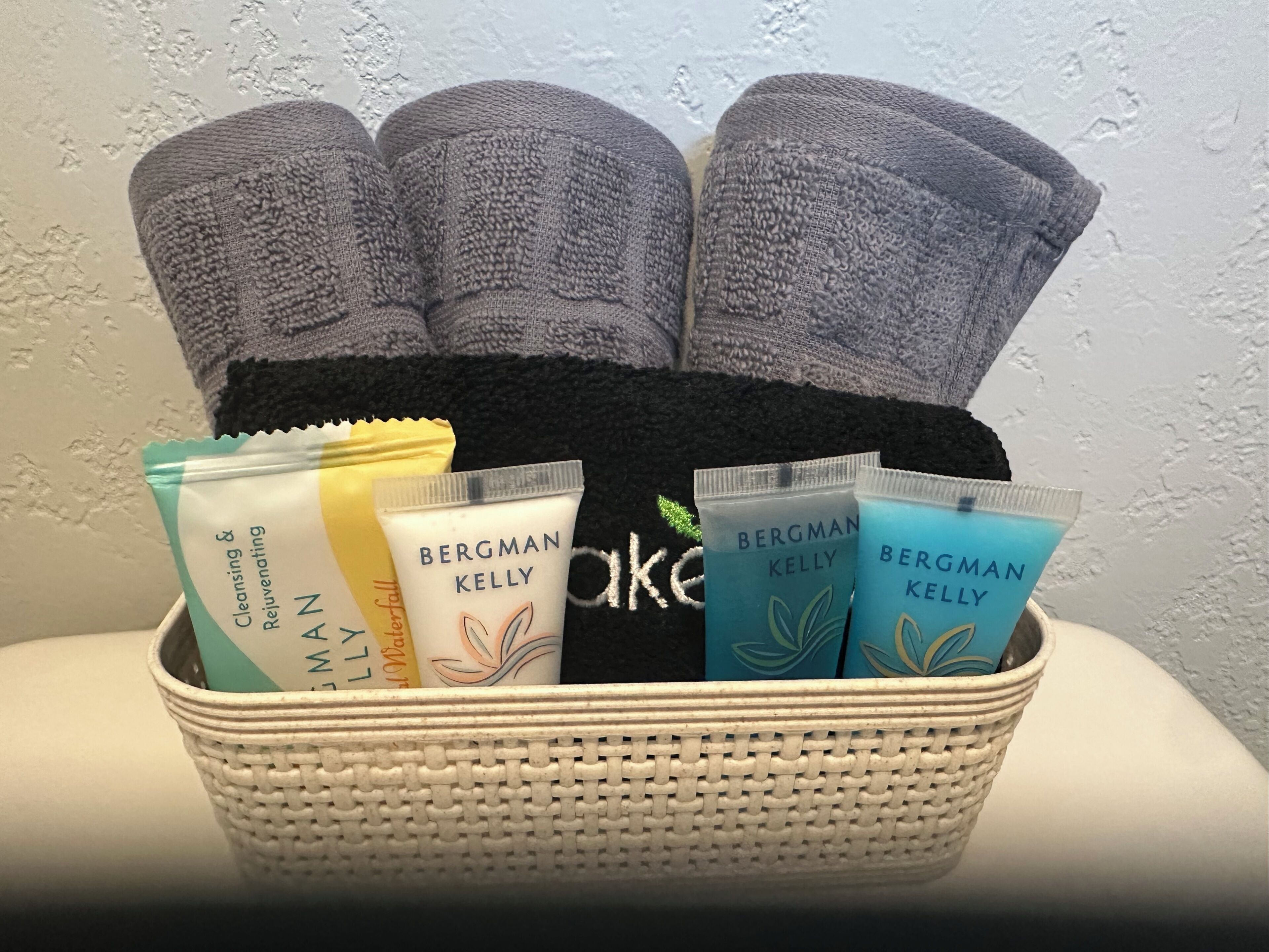 A basket containing towels, a black bag, and Bergman Kelly skincare products.