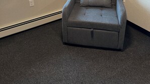 Quarto de canto com poltrona cinzenta, uma janela grande e piso carpeteado.