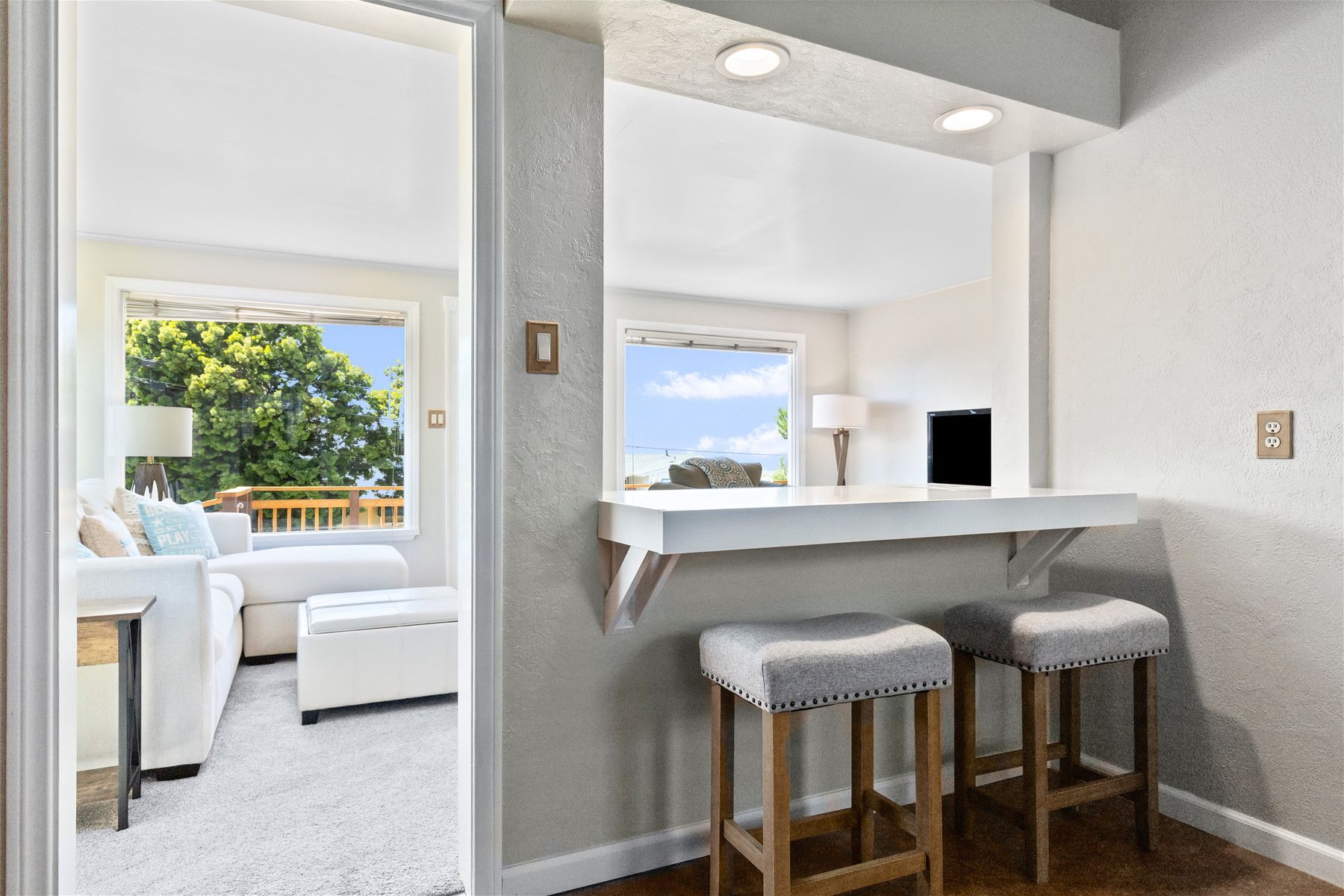 A modern interior with a white ceiling, recessed lighting, a white counter with stools, and a view of greenery outside.