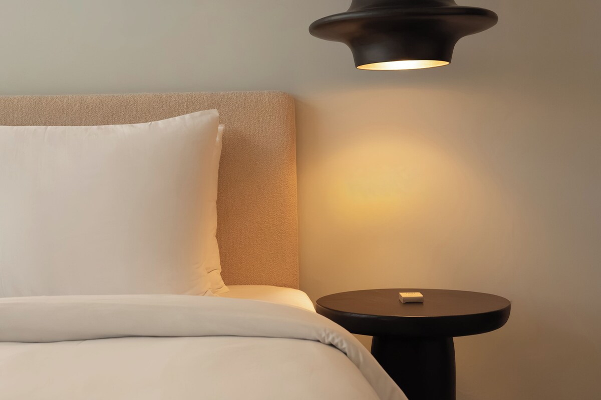 Una cama con ropa de cama blanca, una mesita redonda y una lámpara colgante.