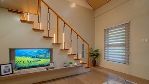 A modern living room with a wooden staircase, a flat-screen TV, and a large window with blinds. - Ohhana Lofts El Nido (El Nido)