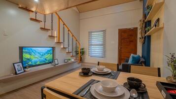 Una sala de estar moderna con una escalera de madera, televisor de pantalla plana, mesa de comedor dispuesta con fuentes, y una zona de estar acogedora con un cojín azul.