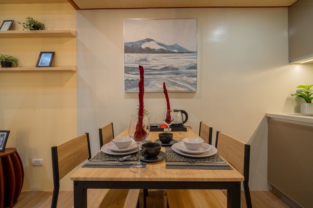 A dining table set with plates, bowls, and glasses, flanked by chairs, with a painting above and a shelf with items on the wall.