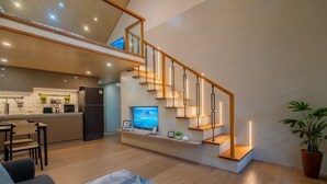 Eine moderne Küche mit integriertem Kühlschrank, eine Essecke mit Tisch und Stühlen, eine Treppe mit integrierter Beleuchtung sowie ein Wohnzimmer mit Sofa und großem Teppich.