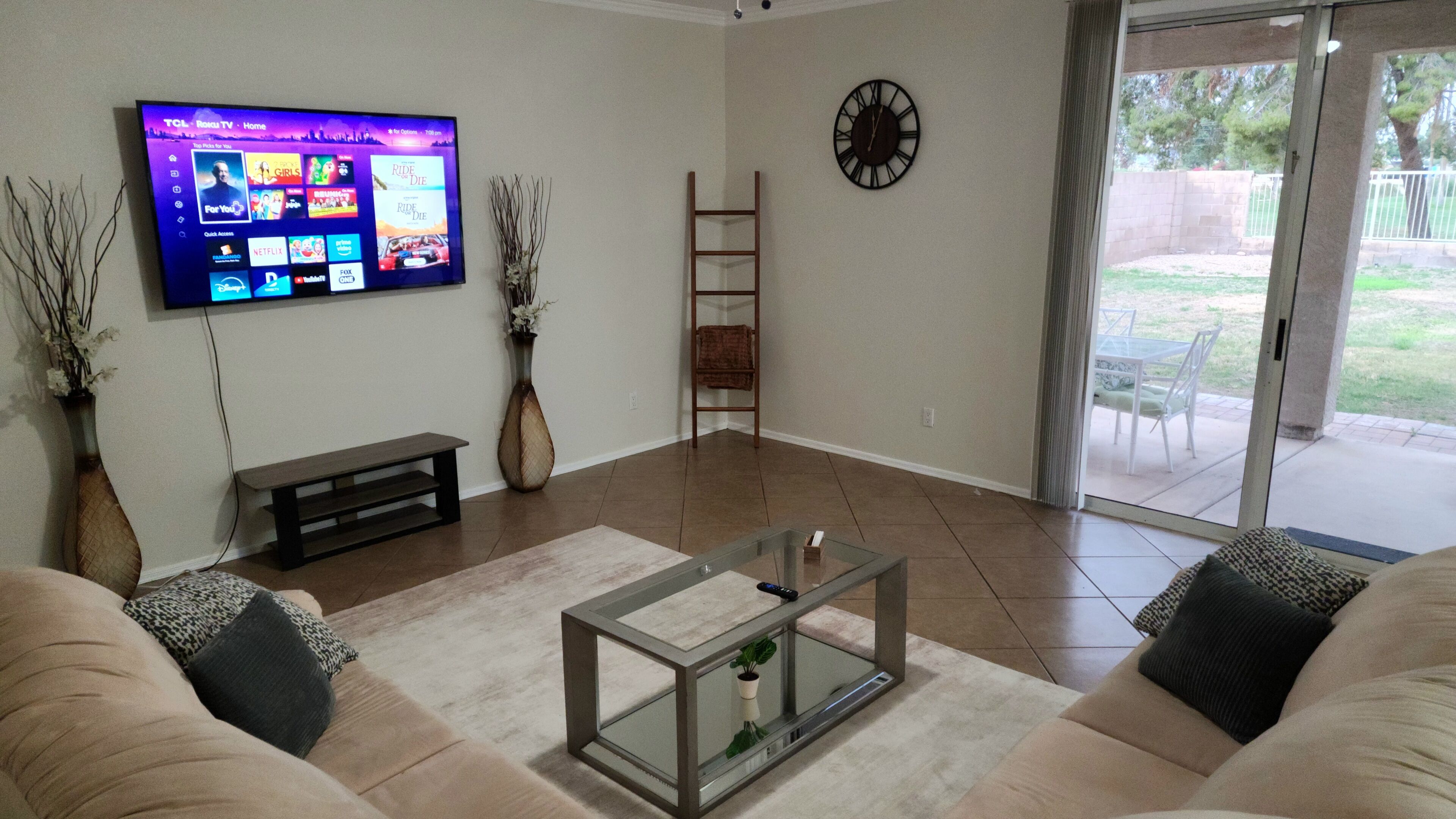 A living room with a flat-screen TV, a bench, a glass coffee table, a clock, and a sliding glass door leading to an outdoor area.