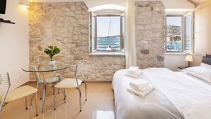 Habitación de hotel con pared de piedra, cama con ropa blanca, mesa de comedor con sillas y vista al mar a través de una ventana.