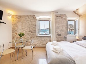Habitación de hotel con pared de piedra, cama con ropa blanca, mesa de comedor con sillas y vista al mar a través de una ventana.