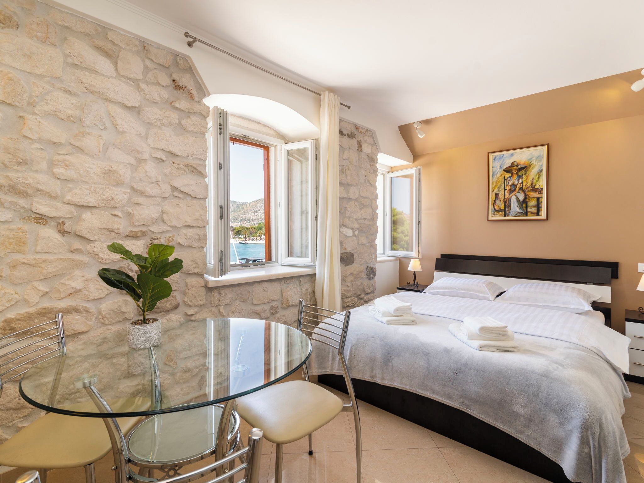 Habitación de hotel con pared de piedra, cama con ropa de cama blanca, una mesa de vidrio con sillas y un cuadro en la pared.