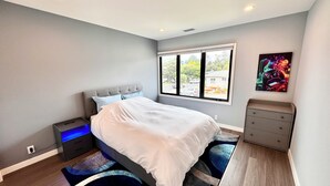 Ein modernes Schlafzimmer mit Bett, Nachttisch, Kommode, einem Fenster mit Ausblick und einem Wandbild.