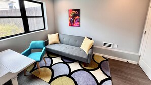 Ein Zimmer mit grauem Sofa, blauem Sessel und weißem Tisch. Ein farbenfrohes Kunstwerk ziert die Wand, und ein Fenster bietet Blick auf einen Zaun.