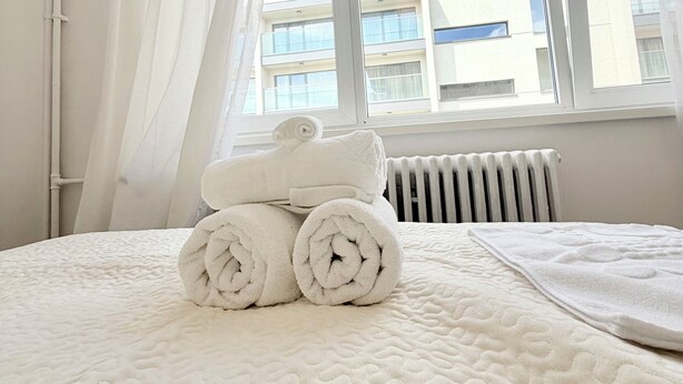 A neatly made bed with rolled towels, a radiator, and a window with curtains. - Limons Apartment: Living Room and Bedroom in Downtown Bucharest (București)