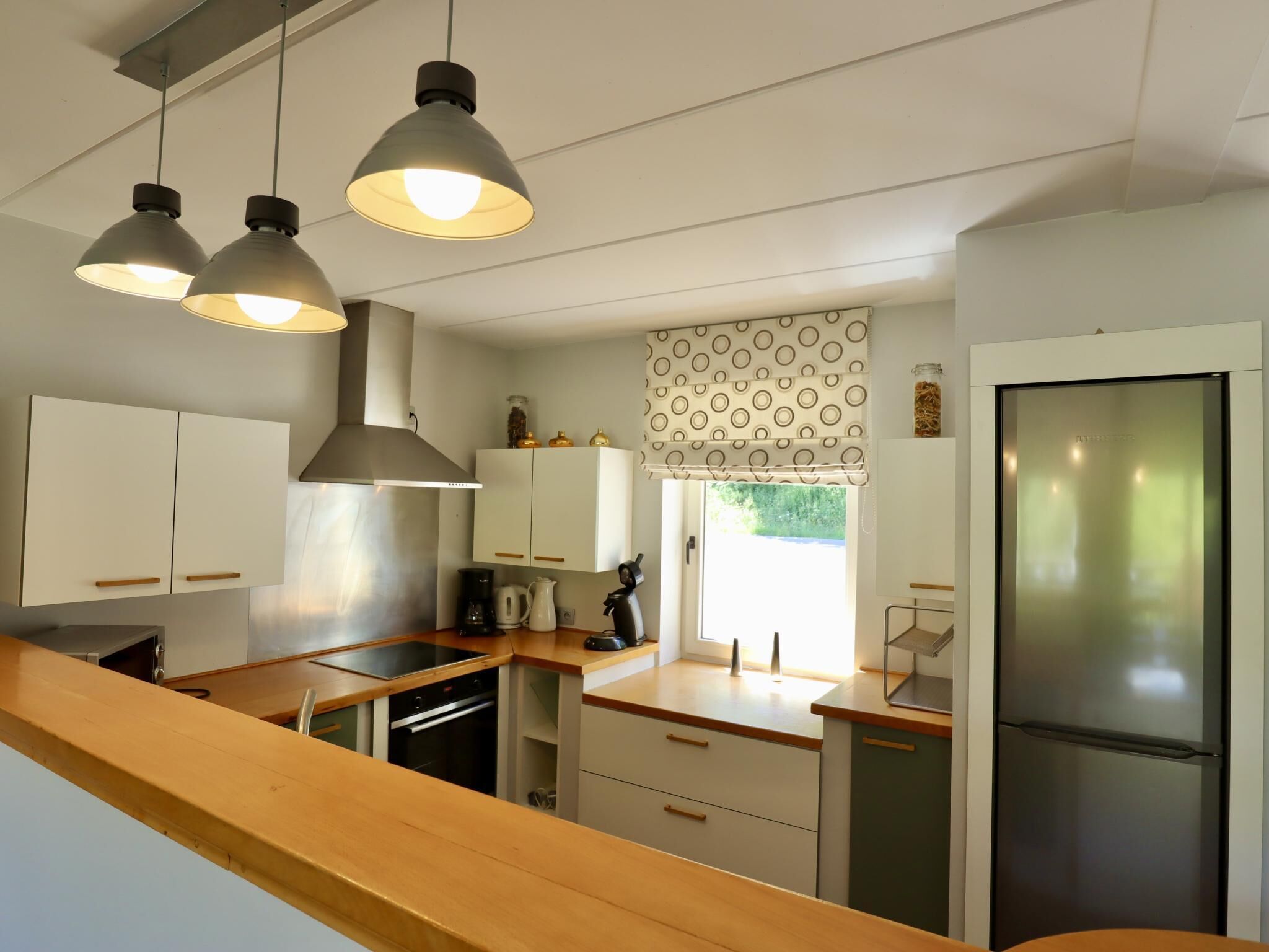 Une cuisine moderne dotée d’un éclairage suspendu, d’un réfrigérateur et d’une fenêtre avec un store à motifs.