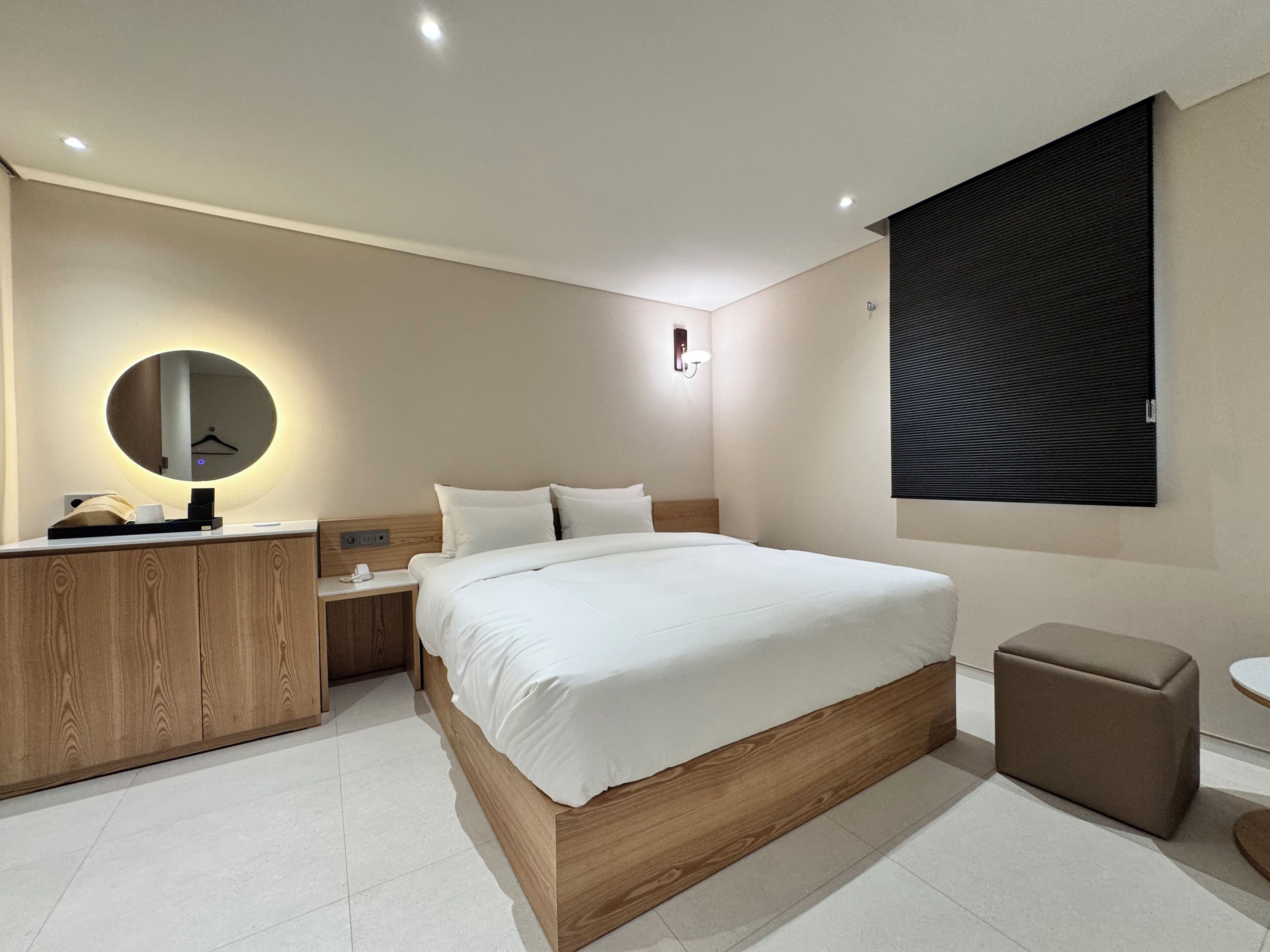 A modern hotel room with a wooden bed, a round mirror, a bedside table with a lamp, a small stool, and a large window with blinds.