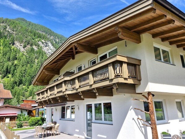 Un bâtiment à deux étages doté d’un balcon, avec une chaîne de montagnes en arrière-plan et un patio aménagé avec du mobilier d’extérieur.