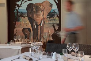 A dining area with tables set for a meal, a painting of an elephant on the wall, and a person walking by. - hôtel les 3 fontaines (Marconne)