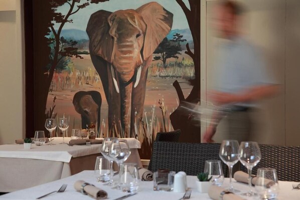 A dining area with tables set for a meal, a painting of an elephant on the wall, and a person walking by.