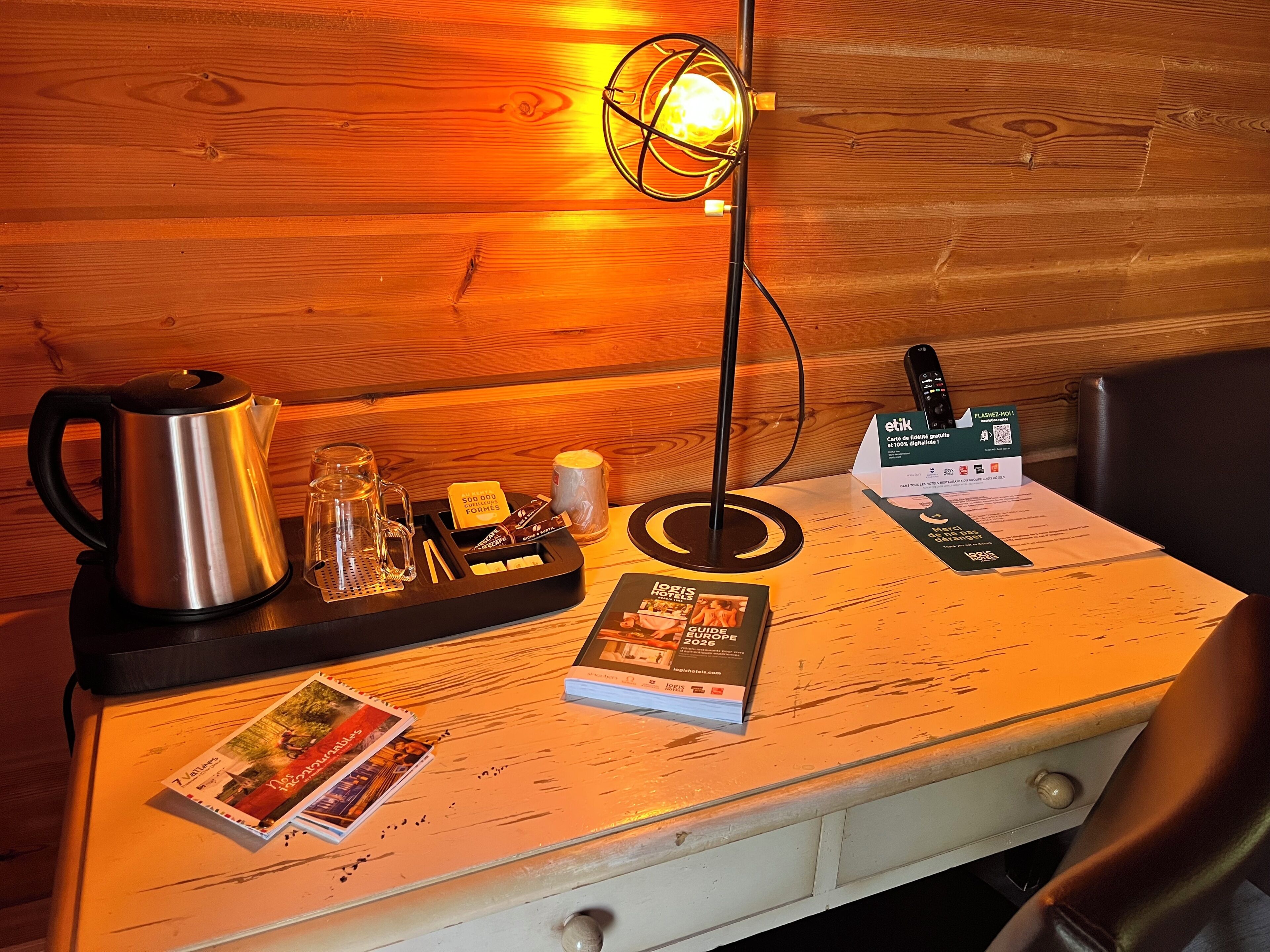 A wooden table with a kettle, a glass, a mug, a remote control, and a guidebook.