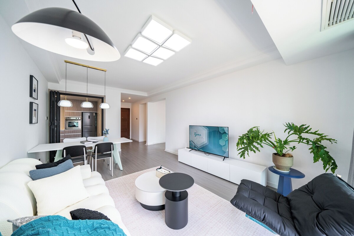 A modern living room with a sofa, a coffee table, a TV, a dining area, and a kitchen in the background.