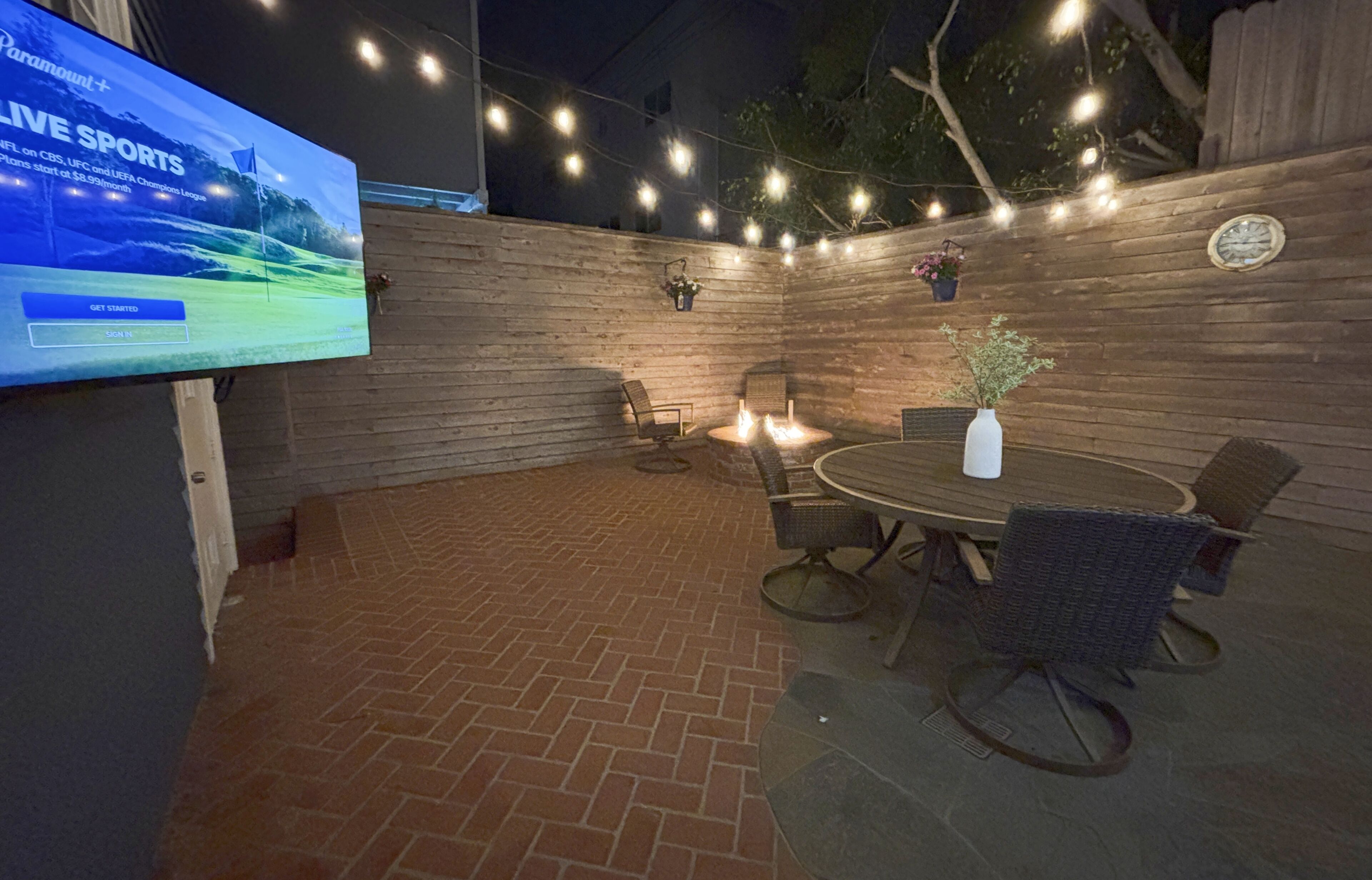 An outdoor patio with a brick floor, a round table with chairs, string lights, and a mounted television displaying sports content.