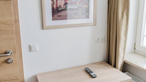 A room with a wooden desk, a remote control, a framed picture on the wall, a window with curtains, and a radiator.