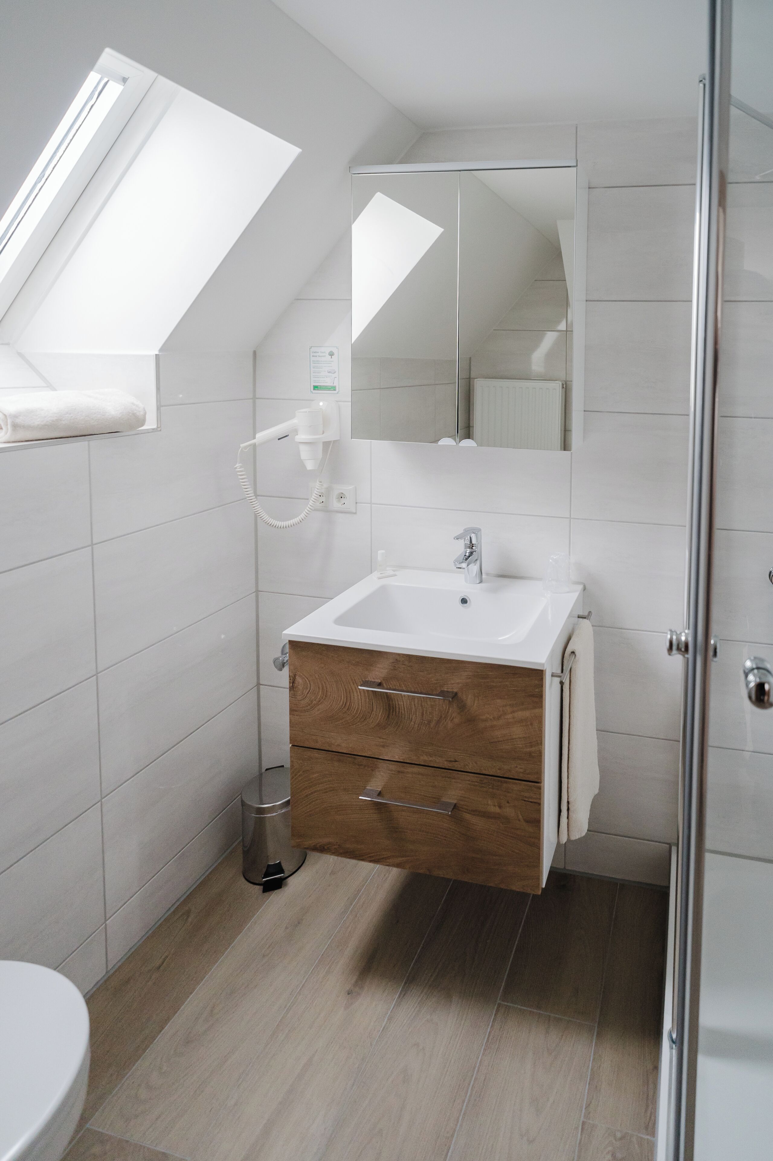A modern bathroom with a skylight, white tiles, a wooden vanity, and a glass shower enclosure.