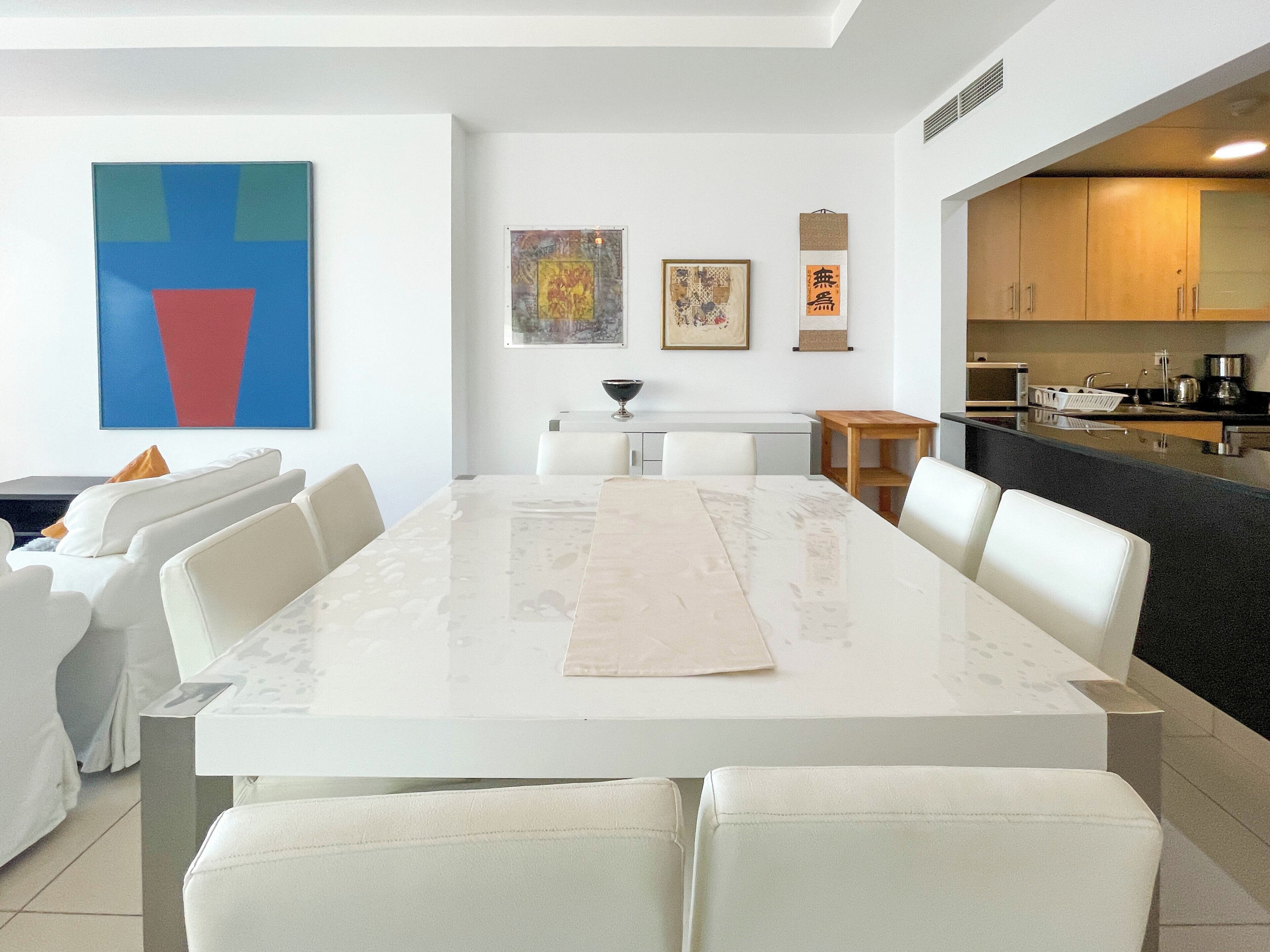 Une cuisine moderne avec un coin repas, des meubles blancs et des œuvres d’art accrochées aux murs.
