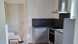 Eine kleine Küche mit Herd, Backofen und kleinem Kühlschrank. Ein Holzhocker und ein weißer Tisch sind ebenfalls vorhanden.