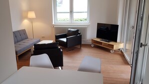 Ein modernes Wohnzimmer mit einer Couch, einem schwarzen Sessel, einem Fernseher auf einem Holzgestell und einem Fenster mit Blick.
