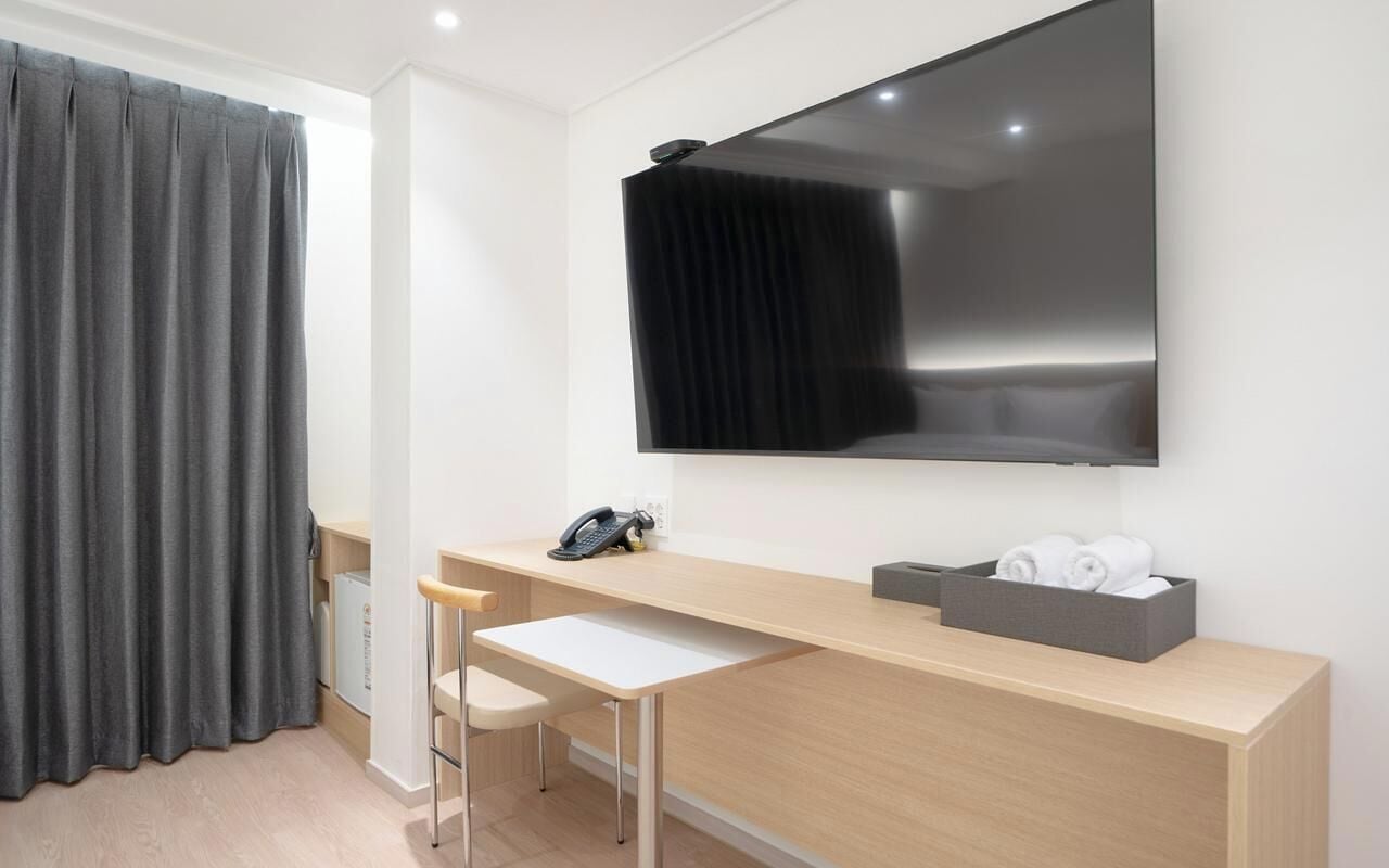 A modern hotel room with a large flat-screen TV mounted on the wall, a wooden desk with a chair, a telephone, and a basket of towels.
