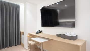 A modern hotel room with a large flat-screen TV mounted on the wall, a wooden desk with a chair, a telephone, and a basket of towels.