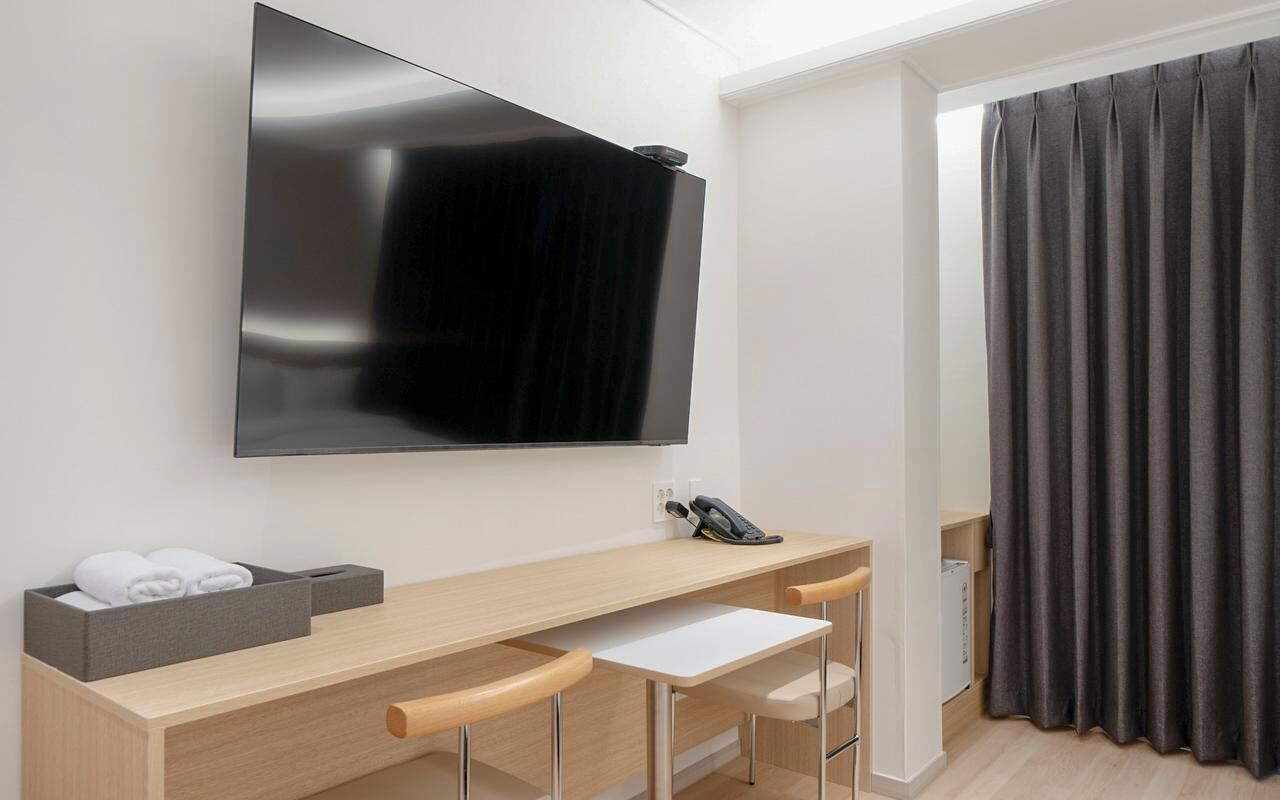 A hotel room with a flat-screen TV mounted on the wall, a desk with two chairs, a telephone, and a dark curtain.