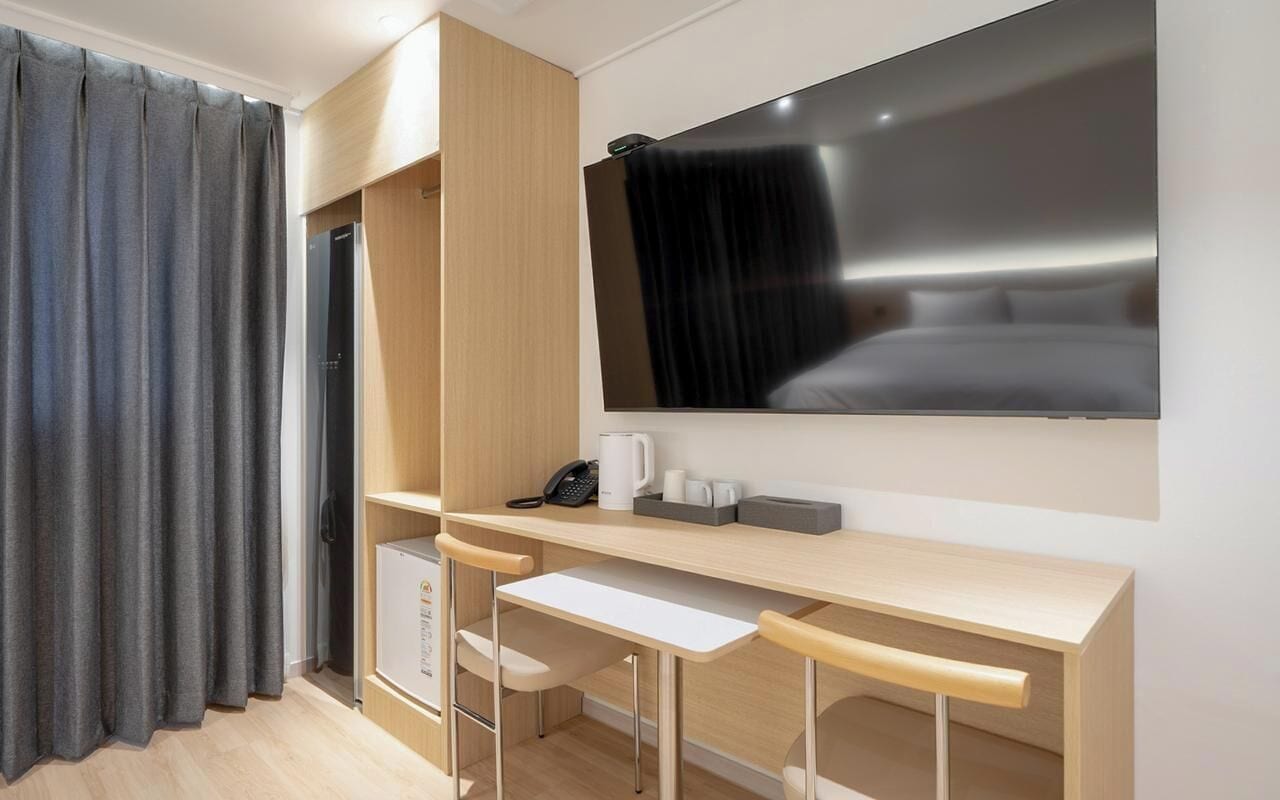 A modern hotel room with a large flat-screen TV, a desk with two chairs, a coffee maker, and a telephone.
