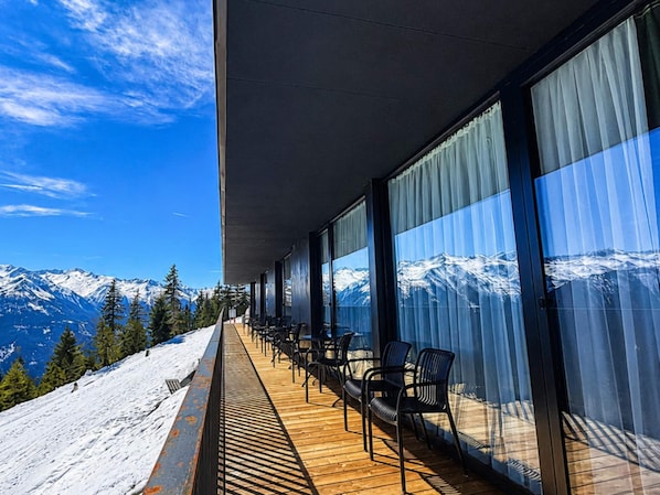Ein Balkon mit Blick auf schneebedeckte Berge, Holzboden und schwarzen Stühlen.