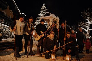 Eine Gruppe von Personen in Winterkleidung hat sich um eine Konstruktion mit weißem Dach, möglicherweise eine Laube, in einer verschneiten Landschaft versammelt. Einige halten Musikinstrumente.