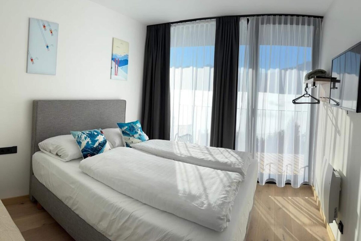 A modern hotel room with a curved bed, white linens, and a large window with sheer curtains.