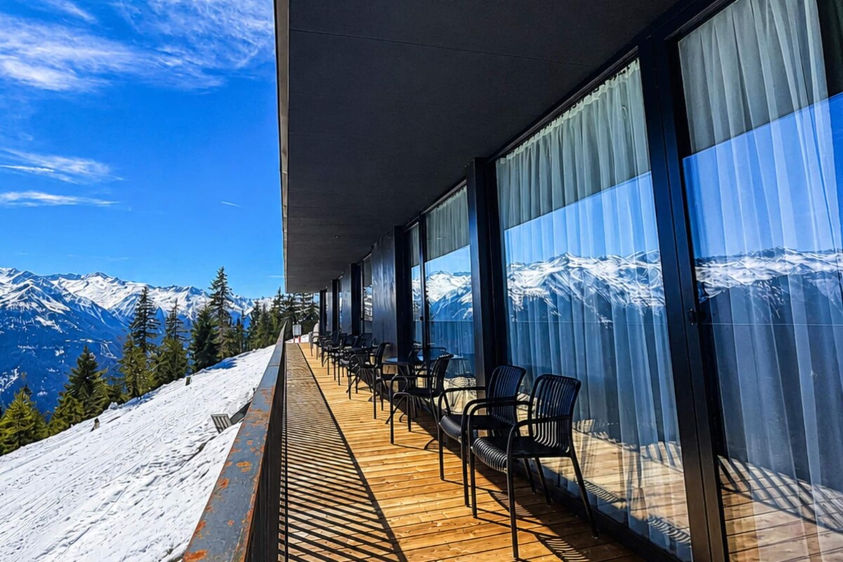 A balcony with a view of snow-capped mountains, wooden flooring, and black chairs.