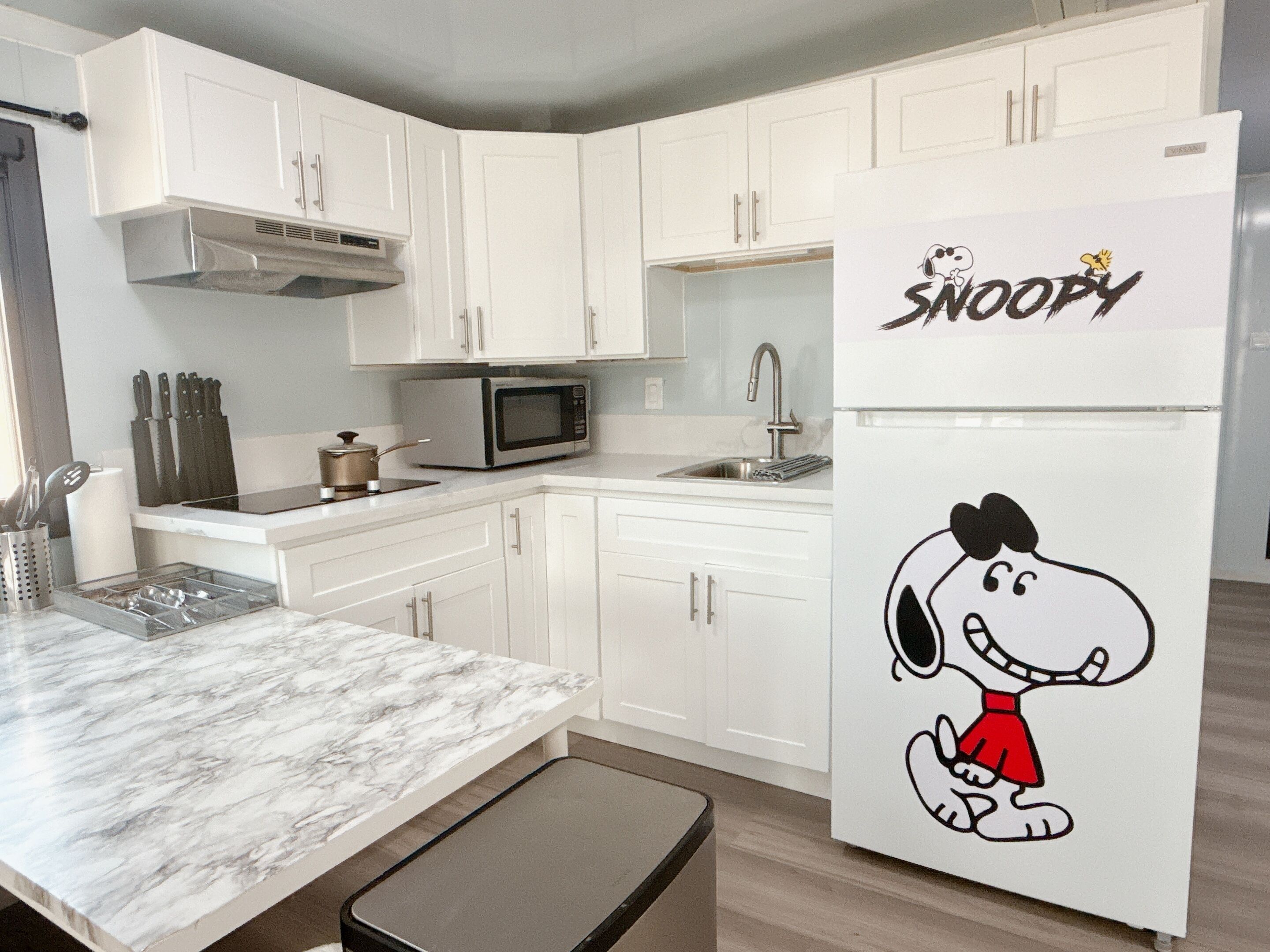 Een keuken met witte kasten, een magnetron, een spoelbak en een Snoopy-koelkast.