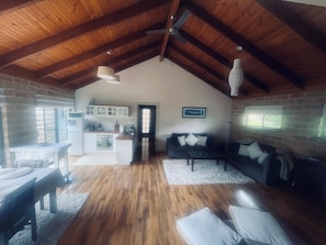 A spacious loft with a high ceiling, exposed wooden beams, a kitchen area, a living room with a sofa and cushions, and a dining area with a table and chairs.