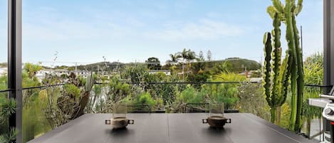 Un balcón con mesa y sillas, con vista a un jardín con diversas plantas y una lejana vista de un paisaje montañoso.