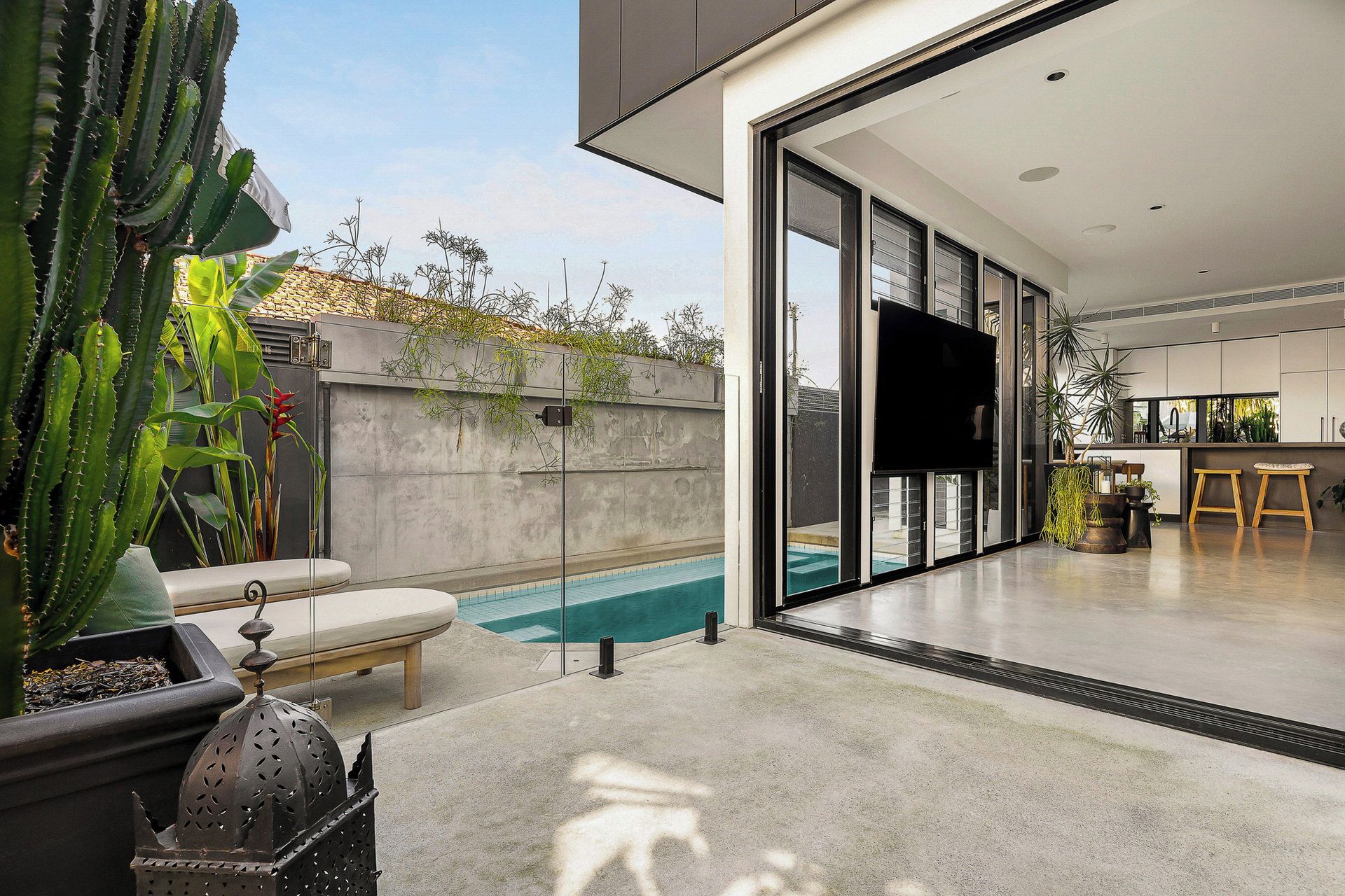 A modern house with a swimming pool, a patio with a lounge chair, and a kitchen area visible through glass doors.